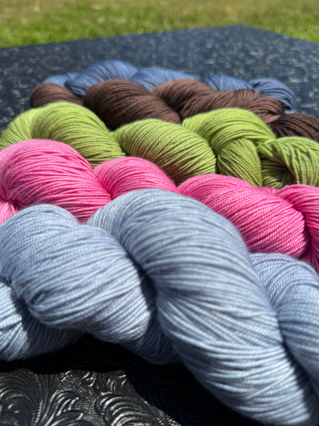 A close-up arrangement of multiple skeins of yarn in different colors, including light blue, pink, olive green, brown, and slate blue, placed on a dark textured surface outdoors with grass visible in the background.